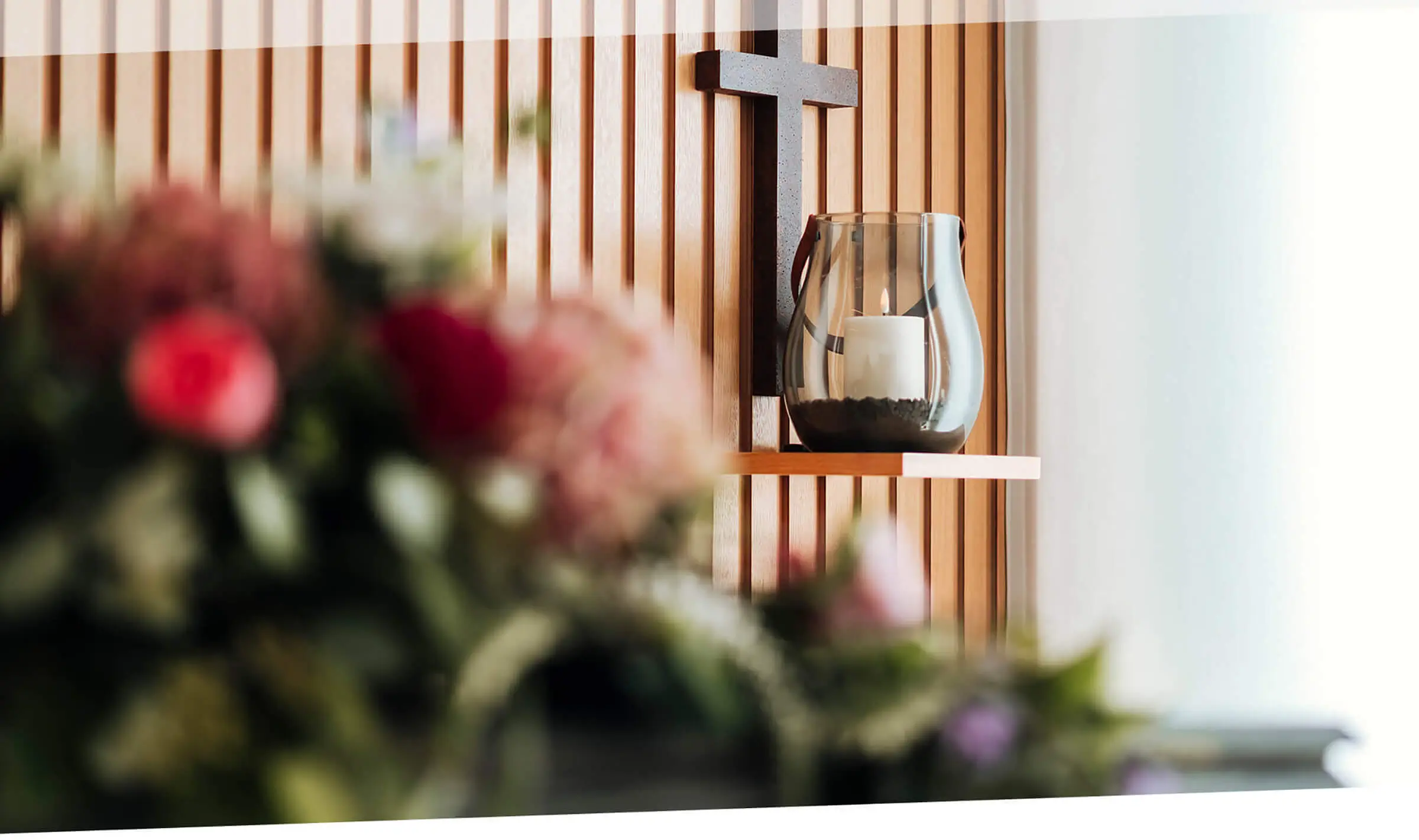 image-raum-4 Ein Kreuz hängt an der Wand. Davor befindet sich auf ein Windlicht mit einer brennenden Kerze. Im Vordergrund sind Blumen zu sehen.